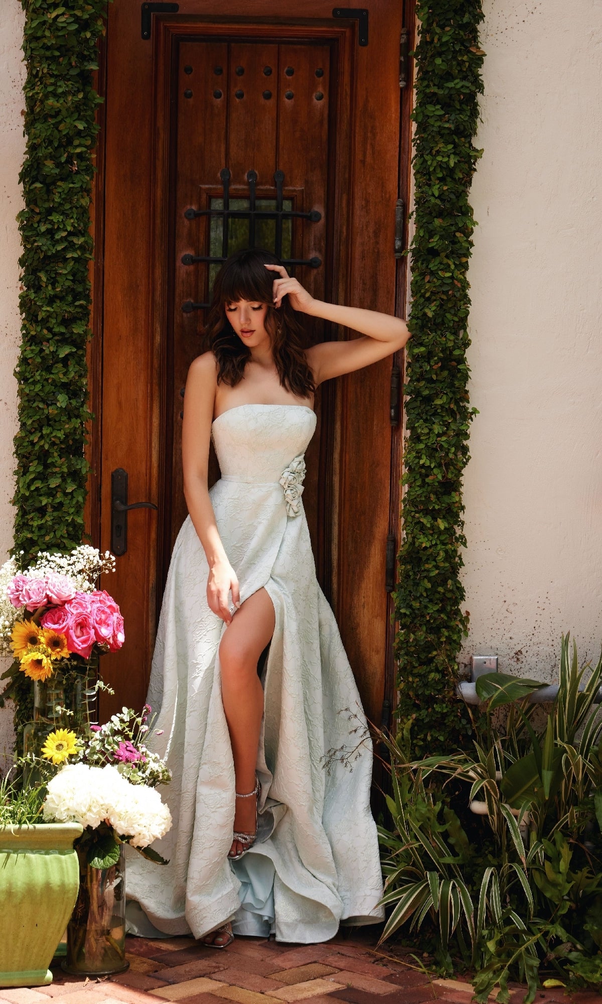 A woman wearing the Rosette A-Line Prom Dress: Ava Presley 40097, featuring a strapless design and high slit, stands by a wooden door framed with greenery and potted flowers.